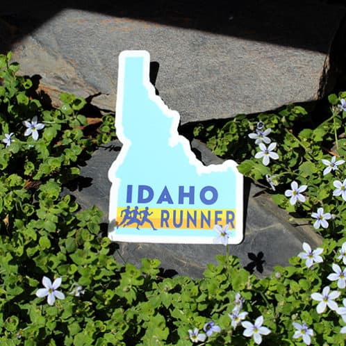 Idaho Running Sticker laying on rocks with white flowers for website