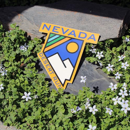 Nevada Running Sticker laying on rocks with white flowers for website