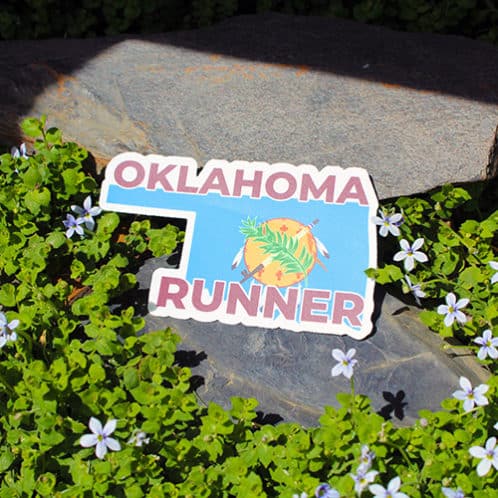 Oklahoma Runner Sticker laying on rocks with white flowers for website
